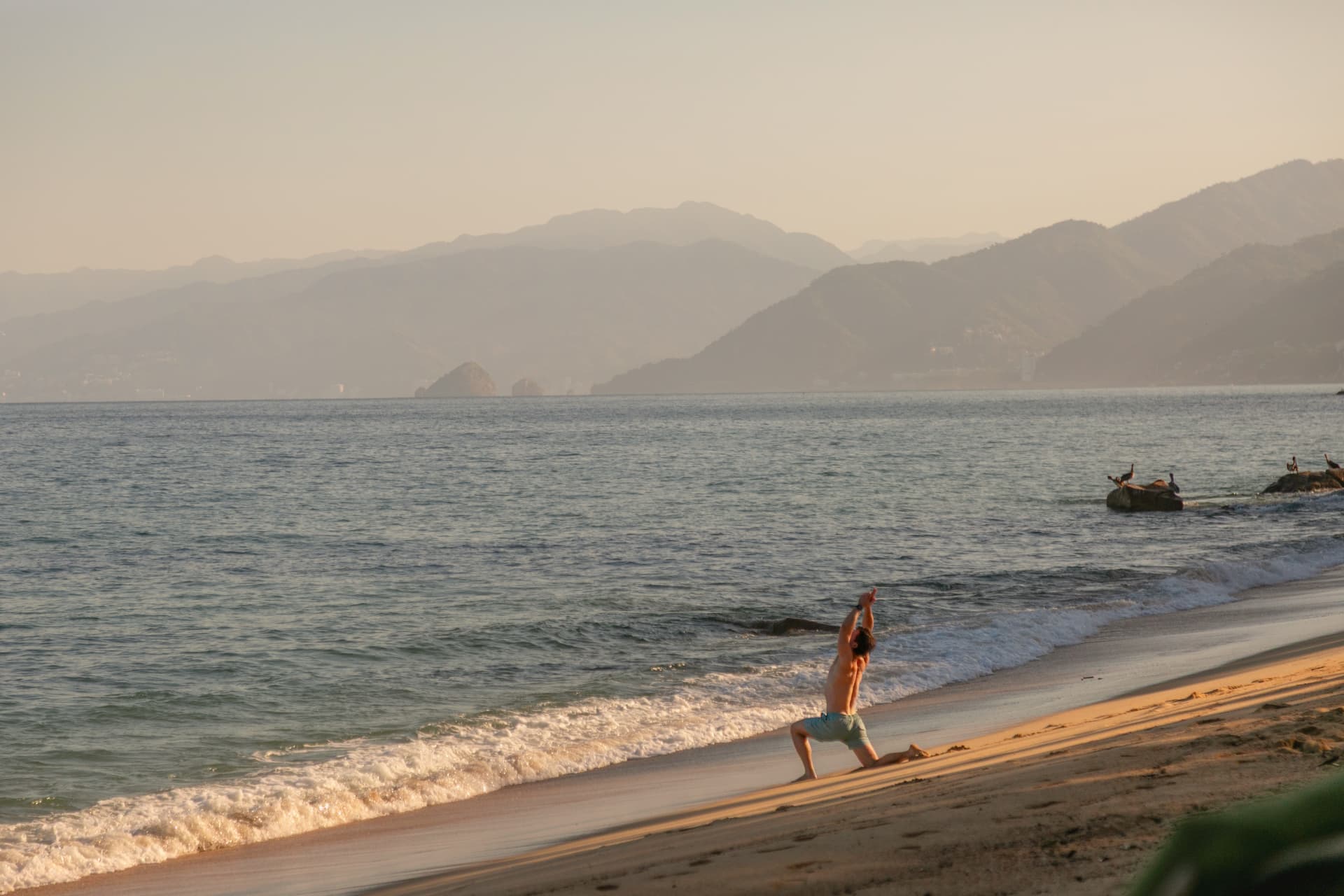 Man practicing yoga at the shoreline at sunrise with the mountains behind