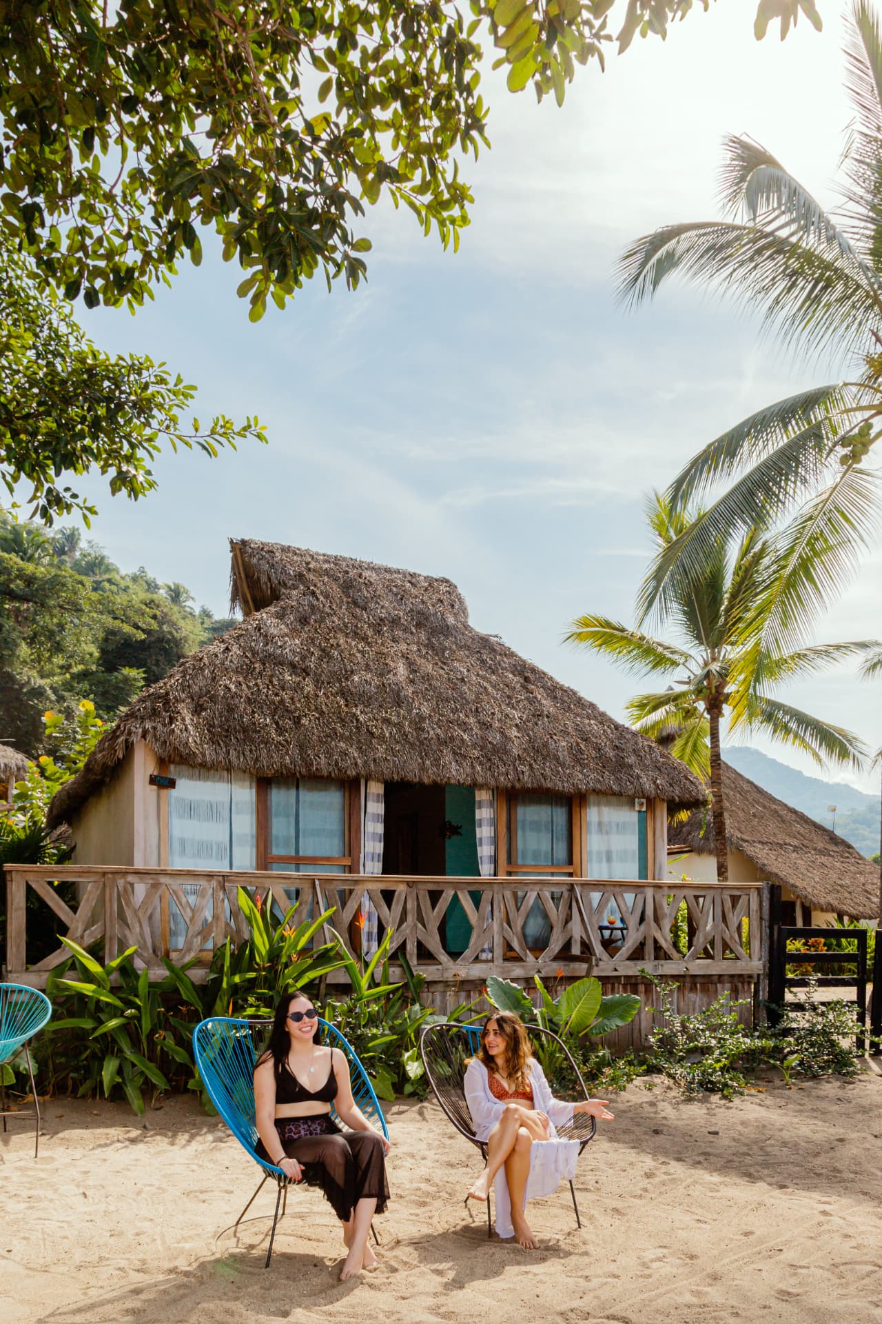 Two friends sitting in Acapulco chairs in front of a palm-roofed cabin