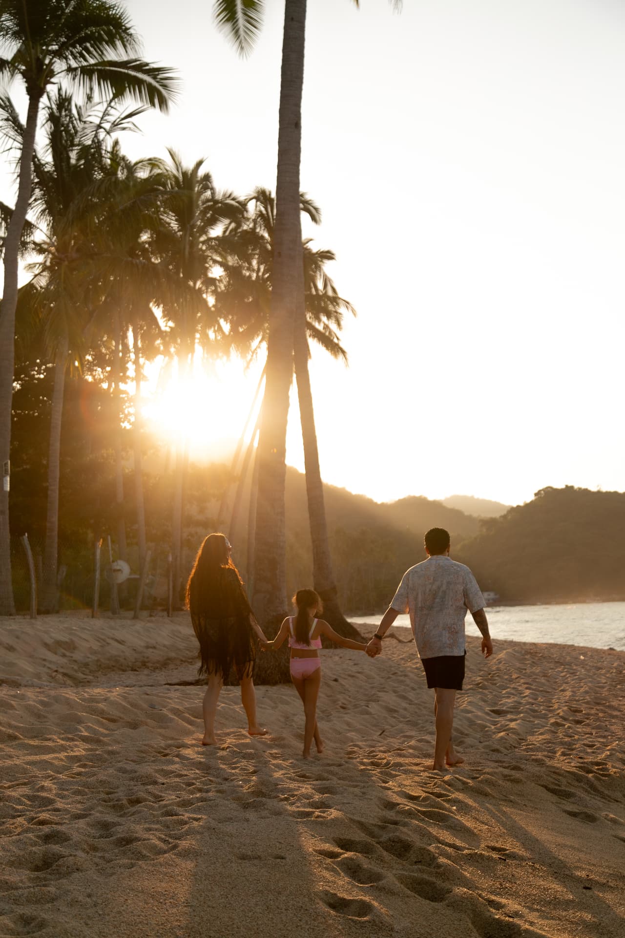 Family walking hand in hand toward the sea among the palms at sunset