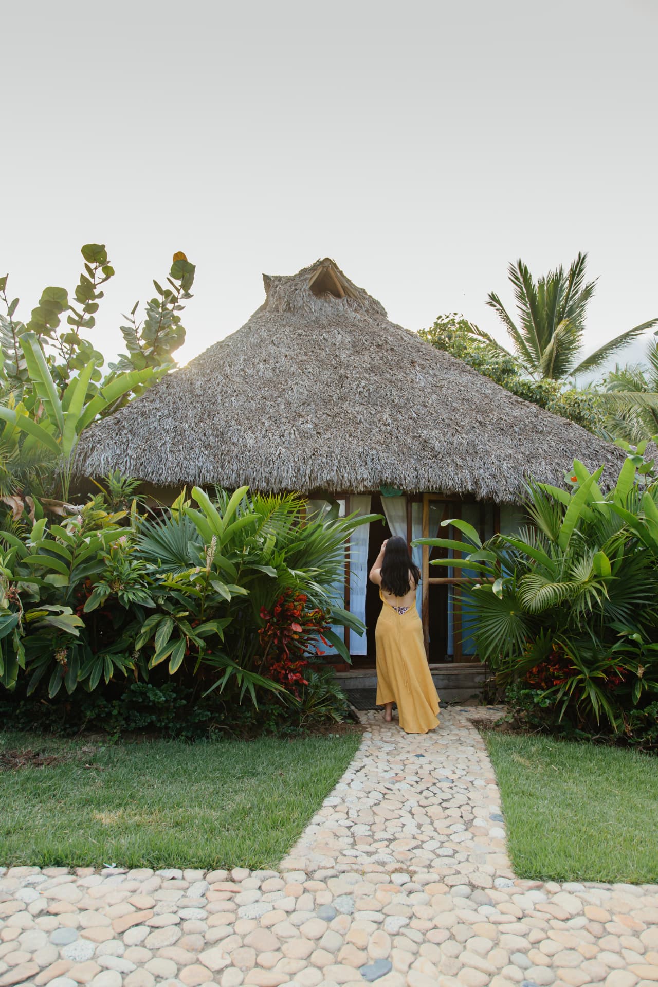 Woman walking into a palm-roofed bungalow surrounded by tropical garden