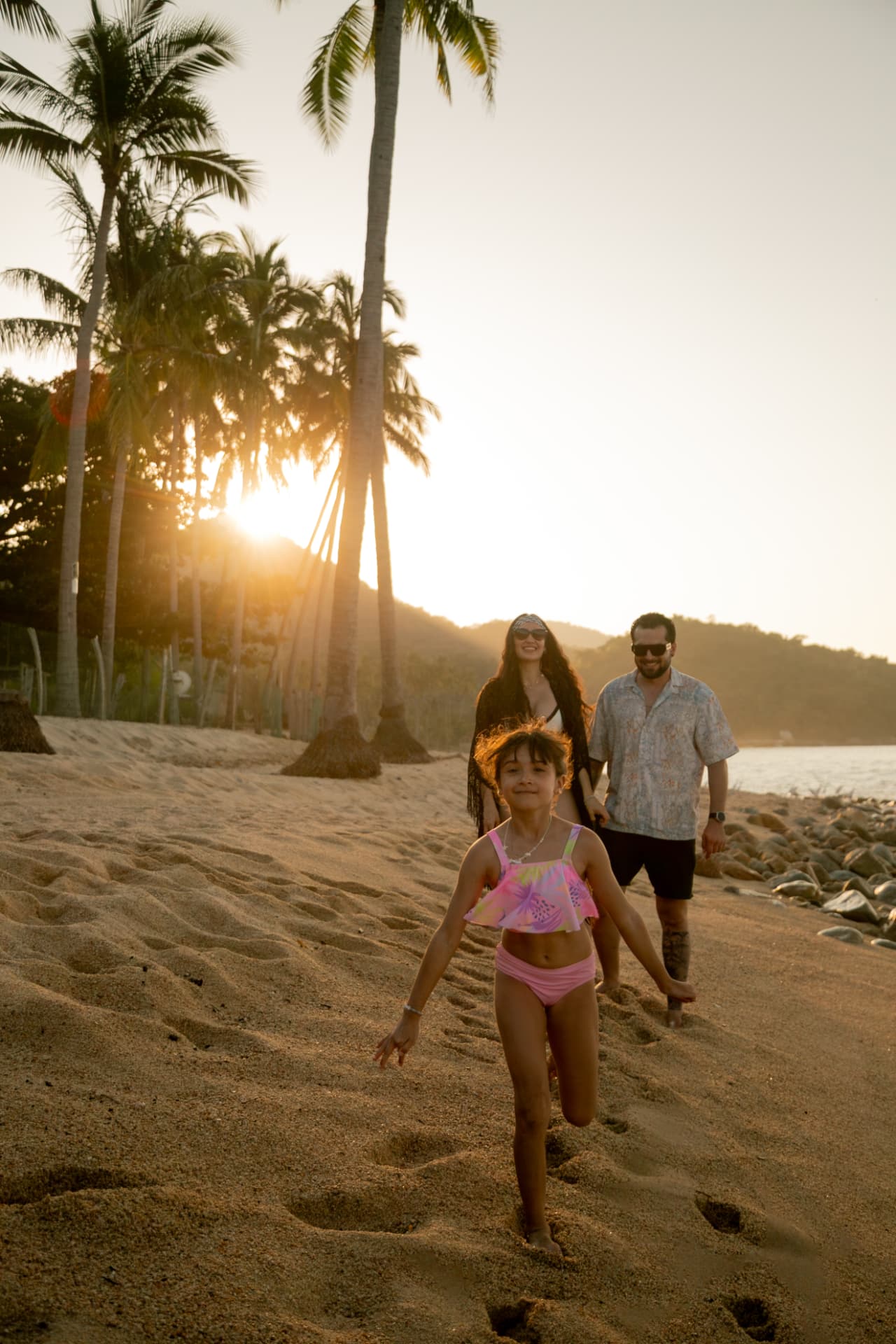 Girl running on the sand followed by her parents under the palms at sunset